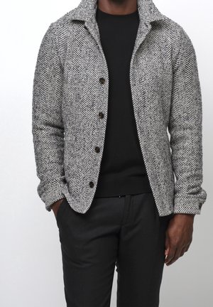 Homme portant une veste boutonnée gris à motif chevron sur une chemise noire et un pantalon noir, les mains dans les poches.