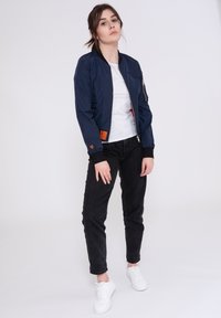 Giubbotto bomber blu notte con zip frontale e due tasche laterali. T-shirt bianca sotto, jeans neri e sneakers bianche. Tessuto morbido e liscio.