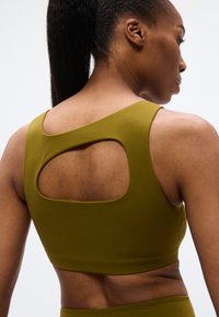 Αθλητικό σουτιέν ελιάς με φαρδιές τιράντες, υφή υφάσματος και σκίσιμο πίσω μέρος, σχεδιασμένο για άνεση και κινητικότητα.