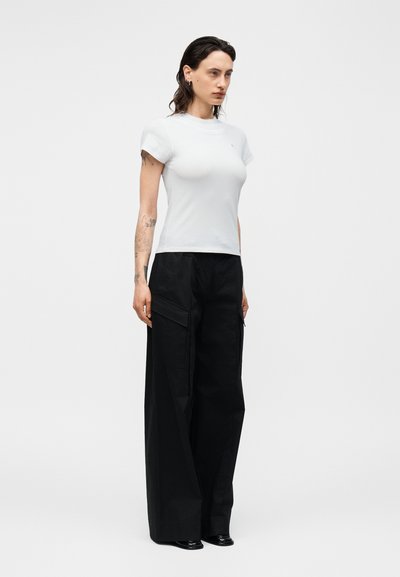 Femme aux bras tatoués portant un haut blanc ajusté à manches courtes et un pantalon noir à jambes larges avec poches latérales, debout devant un fond uni.