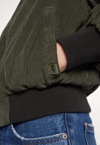 Main dans la poche d'une veste en velours côtelé verte avec un poignet côtelé noir, au-dessus de la poche d'un jean denim bleu avec des coutures visibles et un rivet.