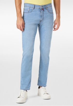 Hombre vistiendo jeans ajustados de color azul claro y zapatillas blancas, de pie contra un fondo blanco liso.