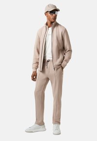 Mand iført beige træningsdragt med lynlåset jakke, matchende bukser, hvide sneakers, beige kasket og overdimensionerede mørke solbriller, der står foran en hvid baggrund.