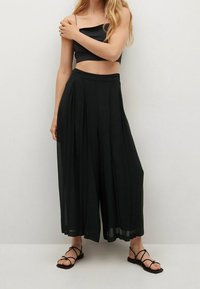 Pantalones anchos de pierna amplia plisados en negro, confeccionados en tejido ligero, con una cintura alta y una silueta fluida, combinados con sandalias negras de tiras.