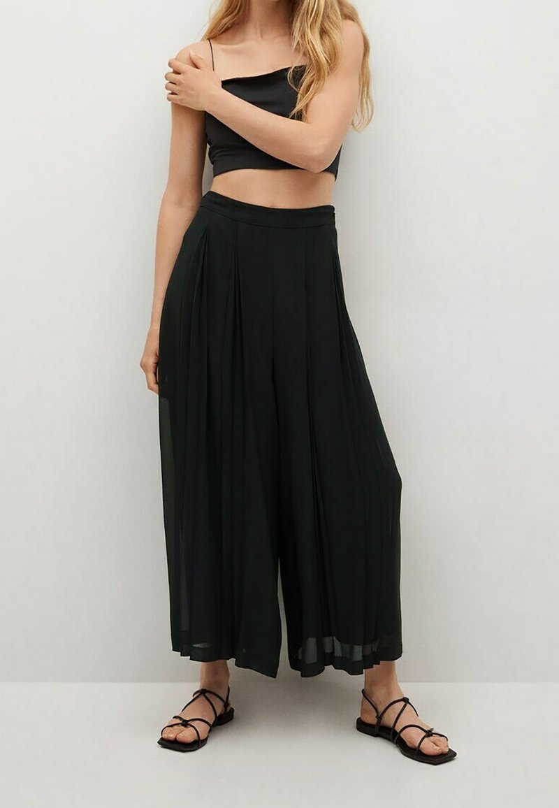 Pantalones anchos de pierna amplia plisados en negro, confeccionados en tejido ligero, con una cintura alta y una silueta fluida, combinados con sandalias negras de tiras.