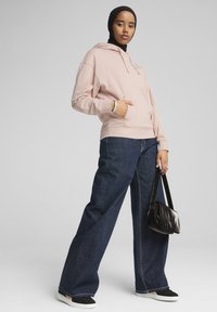 Sudadera con capucha de color rosa claro con bolsillo frontal, combinada con pantalones vaqueros de pierna ancha de color azul oscuro, bolso negro y zapatos negros sin cordones.