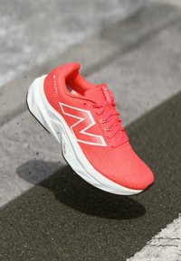 Bright coral New Balance running shoe mid-air above asphalt with white sole and "FuelCell" text on side.