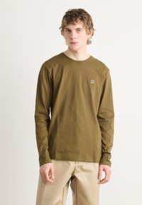 Langarmhemd aus olivgrünem Baumwollstoff mit Rundhalsausschnitt und einem kleinen quadratischen Logopatch auf der Brust. Glatte Textur, lockere Passform.