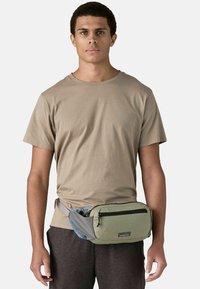 Riñonera en verde y gris con un bolsillo frontal con cremallera, llevada en la cintura. Hecha de tela duradera con una textura suave.