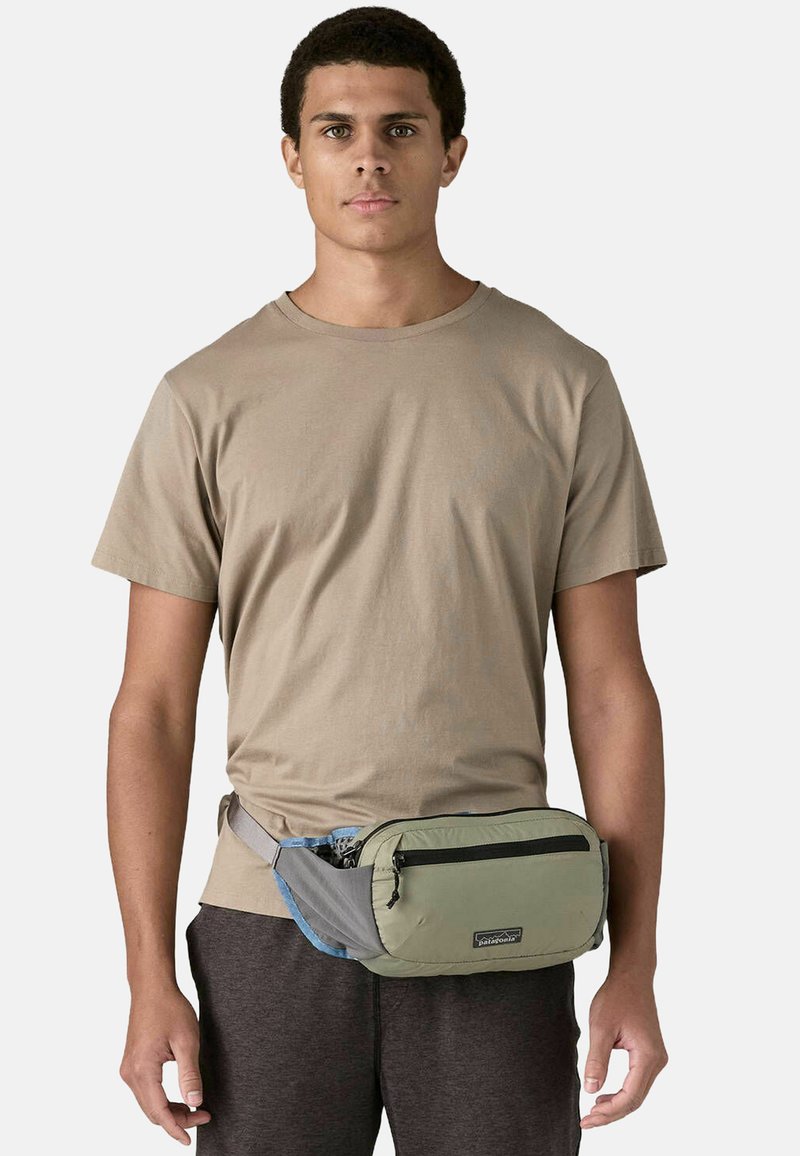 Riñonera en verde y gris con un bolsillo frontal con cremallera, llevada en la cintura. Hecha de tela duradera con una textura suave.