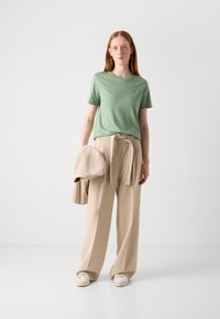 T-shirt verde a maniche corte, pantaloni beige a vita alta con dettaglio in vita legato, in mano un blazer beige coordinato, abbinato a sneakers bianche.