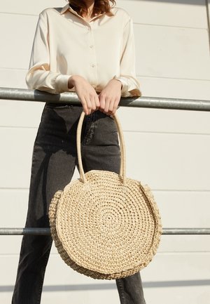 Mujer con blusa color crema y jeans negros sosteniendo un bolso redondo de paja tejido frente a una pared blanca y una barandilla de metal.