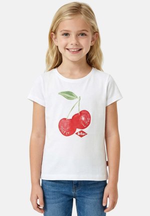 Jeune fille souriante aux cheveux blonds portant un t-shirt blanc avec deux cerises rouges et le texte « Sister Love » ainsi qu'un jean bleu.