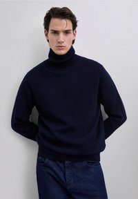 Pull à col roulé bleu marine en maille douce et texturée. Coupe ample avec poignets et ourlet côtelés, assorti à un jean denim foncé.