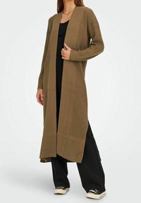 Cardigan long couleur olive avec une texture côtelée, à l'avant ouvert et doté de fentes latérales, porté sur une tenue noire et accentué par des manches longues.