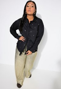 Camisa negra de manga larga con detalles de ojales plateados, combinada con pantalones metálicos dorados de pierna ancha y calzado marrón oscuro. Tejido de textura suave.