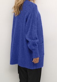 Cardigan de gran tamaño, azul cobalto, hecho de material de punto suave. Presenta puños y dobladillo acanalados, y un bolsillo frontal en el lado izquierdo.