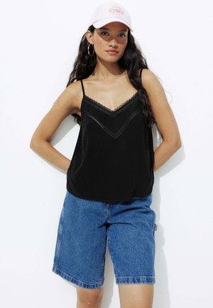 Camisole noire avec bordure en dentelle et fines bretelles, associée à un short en denim bleu clair. Le modèle porte une casquette rose et des boucles d'oreilles créoles.