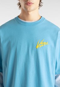 Vaaleansininen puuvillapaita, jossa on pyöreä kaula-aukko, rinnan kohdalla keltainen "Vans"-logo ja sileä pinta.