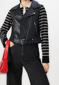 Personne portant un gilet de motard en cuir noir sur une chemise à manches longues rayée noire et blanche, un jean noir, et tenant un sac à main rouge.