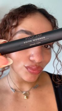 Personne tenant un crayon ombre à paupières GLIDR Hot Sand noir proche du visage, avec des cheveux bouclés, un maquillage naturel et des colliers superposés visibles.