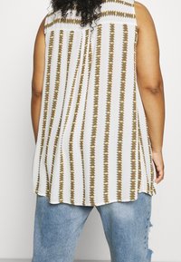 Top senza maniche bianco con motivo a strisce verticali dorate e verdi. Tessuto leggero, vestibilità ampia e orlo curvo. Indossato sopra jeans in denim.