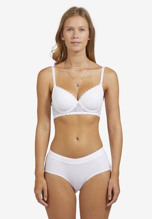 Frau mit hellbraunen Haaren, die einen weißen Spitzen-BH und weiße Baumwoll-Unterhosen trägt, steht vor einem einfarbigen weißen Hintergrund.