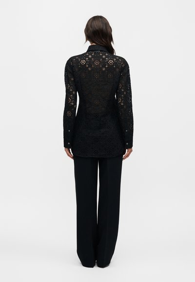 Γυναίκα από πίσω φοράει εφαρμοστό μαύρο δαντελένιο μακρυμάνικο πουκάμισο και φαρδιά μαύρη παντελόνα με φόντο απλό λευκό.