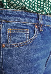 Los jeans de mezclilla azul presentan un pequeño bolsillo frontal, costuras doradas y un remache de cobre. Una tela verde estampada es parcialmente visible en la parte superior.