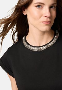 Schwarzes Oberteil mit einem mit Schmuck verzierten Kragen in klaren und roten Akzenten, kurzen Ärmeln und weicher Textur, das eine figurbetonte Silhouette hervorhebt.