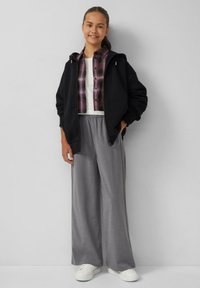 Schwarze Zip-Hoodie über einem lila karierten Hemd, einem weißen T-Shirt und grauen weiten Hosen. Weiße Sneaker vervollständigen das Outfit.