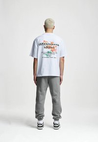 Cămașă albă cu mânecă scurtă, cu grafica florală și textul "VIAȚĂ INTENȚIONATĂ" pe spate, asortată cu pantaloni de trening gri și adidași.