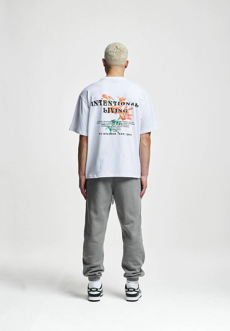 Cămașă albă cu mânecă scurtă, cu grafica florală și textul "VIAȚĂ INTENȚIONATĂ" pe spate, asortată cu pantaloni de trening gri și adidași.