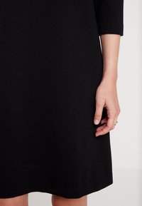 La main droite d'une femme repose à côté d'une robe noire longueur genou à manches trois-quarts sur un fond neutre.
