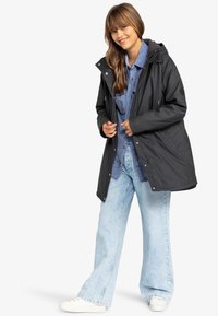 Zwarte waterdichte jas met een capuchon, voor knopen en een trekkoord. Draag het over een blauwe button-up shirt en lichte flared jeans. Witte sneakers.