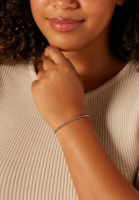 Zilveren armband met kleine kraaldetails, gedragen om een gebruinde arm. Op de achtergrond een geribbelde stof in neutrale kleur.