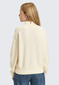 Kremowy sweter z prążkowanym obszyciem na dole i wysokim dekoltem z subtelnym wycięciem z tyłu. Miękka, teksturowana dzianina zapewnia delikatną elastyczność.