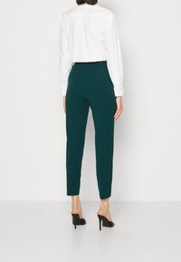 Kvinna iklädd mörkgröna högmidjade byxor, vit långärmad skjorta och svarta högklackade skor, vänd bort från kameran mot en vit bakgrund.