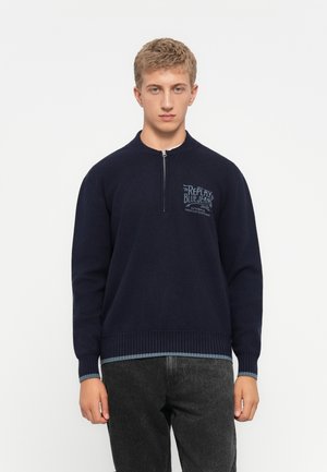 Young man standing, wearing a navy blue quarter-zip sweatshirt with "Replay Blue Jeans" text on chest and black jeans against white background.