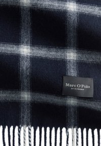 Dunkelblau kariertes Wollbettlaken mit einem weißen Fransenrand, versehen mit einem schwarzen Etikett, auf dem "Marc O'Polo" und "EST. IN STOCKHOLM." steht.