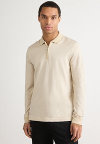 Beige langermet polo skjorte med glidelås-kra, teksturert stoff og tettsittende design, parret med svarte bukser.