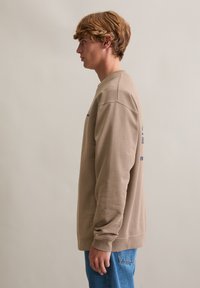 Beżowa bawełniana bluza z okrągłym dekoltem, długimi rękawami i ściągaczami na mankietach. Na lewej stronie znajduje się niebieski napis. Noszona z jasnoniebieskimi dżinsami.