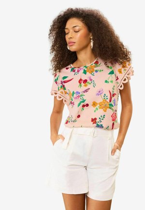 Göndör hajú nő rózsaszín virágos felsőben, amely csipkés ujjú, és magas derekú fehér rövidnadrágot visel, kezét a zsebében tartja, szeme csukva.