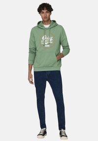 Sudadera verde con cordones, bolsillo frontal y estampado de logo. Combinada con jeans azul oscuro y zapatillas negras con suela crema.