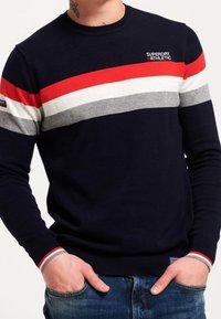 Tmavě modrý svetr s červenými, bílými a šedými vodorovnými pruhy, kulatým výstřihem a žebrovanými manžetami. Na hrudi je logo a kontrastní detaily.