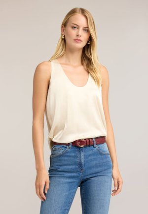 Donna bionda che indossa un top crema senza maniche infilato nei jeans blu con una cintura rossa, in piedi davanti a uno sfondo beige semplice.