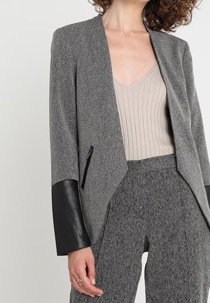 Grå kavaj med texturerad tyg och svarta läderslevar, öppen design, smal passform, tillsammans med en ribbad beige topp och matchande byxor.