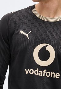 Άνδρας που φοράει μαύρη αθλητική φανέλα με λογότυπο Puma στο αριστερό στήθος και μεγάλο λογότυπο Vodafone κεντραρισμένο κάτω από το γιακά.
