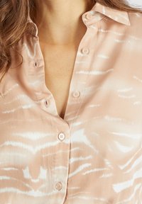 Blusa beige de botones con una textura suave y sedosa, que presenta un sutil patrón abstracto blanco y un cuello clásico.