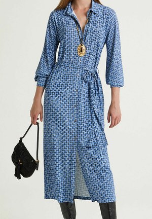 Blue patterned shirt dress with long sleeves, button-down front, waist tie, and side slits; paired with a black handbag.